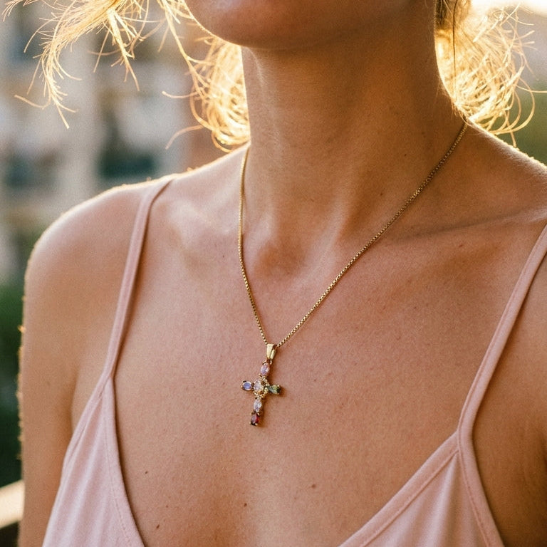 Woman's collarbone wearing gold multicolor stone cross pendant necklace, cross pendant with colored stones catching warm golden hour sunlight, pink slip dress, blurred outdoor background with warm bokeh
