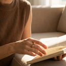 Woman's hand turning book pages wearing gold geometric multi-stone ring with deep green and clear baguette stones on index finger, warm afternoon sunlight, camel ribbed knit vest, blurred beige sofa background
