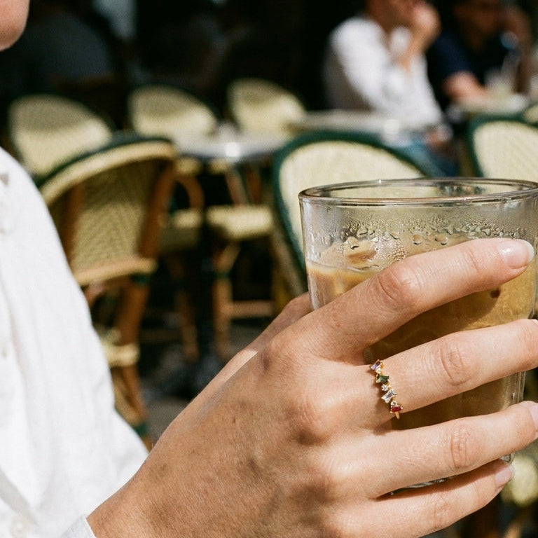 Woman's hand holding glass at outdoor European cafe wearing gold geometric multi-stone ring with multicolor baguette stones on ring finger, white shirt, blurred rattan chairs and cafe crowd in background
