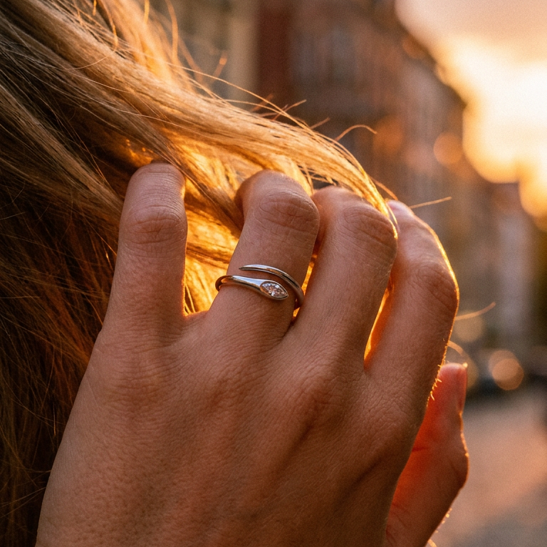 Woman's hand running through hair wearing silver marquise stone snake ring on middle finger, bezel-set marquise stone catching warm golden hour backlight, open band visible, blurred city street and warm sunset bokeh background
