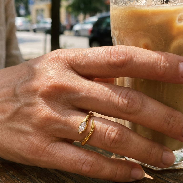 Woman's hand holding iced latte glass at outdoor cafe wearing gold marquise stone snake ring on ring finger, bezel-set marquise stone facing upward, open band gap visible, blurred European street background

