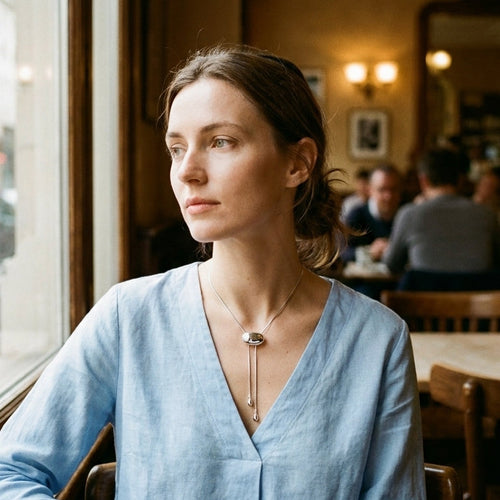 Woman sitting by cafe window wearing gold oval slider lariat necklace, oval slider visible at V-neckline of light blue linen shirt, two chain ends falling below, warm indoor light with blurred cafe background
