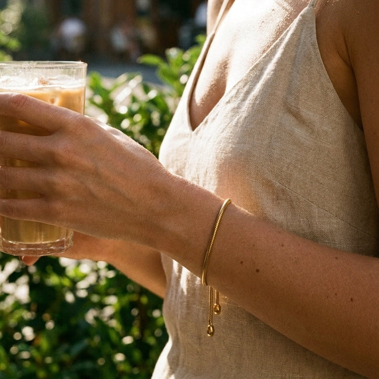 Woman's wrist wearing gold snake chain slider bracelet while holding iced latte glass, fine snake chain lying flat on wrist with two oval tips hanging below, beige linen top, blurred green garden background

