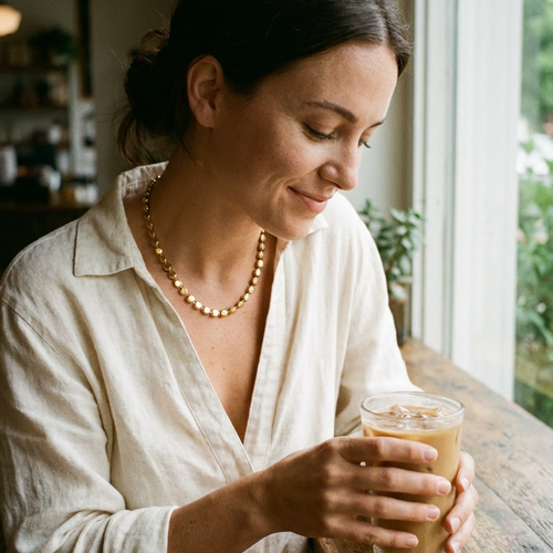 Woman sitting by cafe window holding iced latte wearing gold disc chain necklace, flat disc links visible at collarbone in soft natural window light, white linen shirt, dark hair in bun, blurred indoor cafe background
