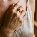Woman's hand resting on chest wearing gold multicolor heart stone open ring on middle finger, pink, purple, green and olive heart-cut stones catching warm indoor light, beige ribbed vest, blurred warm wood background
