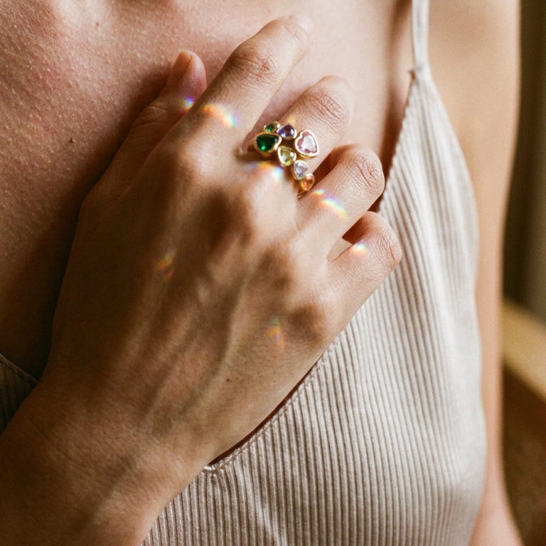 Woman's hand resting on chest wearing gold multicolor heart stone open ring on middle finger, pink, purple, green and olive heart-cut stones catching warm indoor light, beige ribbed vest, blurred warm wood background

