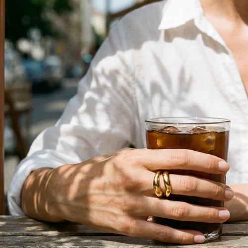 Woman's hand holding iced coffee glass at outdoor cafe wearing gold double wave band ring on middle finger, two-band structure catching bright natural sunlight, white shirt, blurred street background
