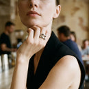 Woman resting chin on hand in indoor cafe wearing silver double wave band ring on index finger, two sculptural bands visible in warm ambient light, black sleeveless V-neck top, blurred cafe crowd in background
