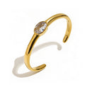 Gold oval stone open cuff bracelet in clear on white background, smooth high-polish band with bezel-set clear oval stone at center, open back visible, long warm shadow
