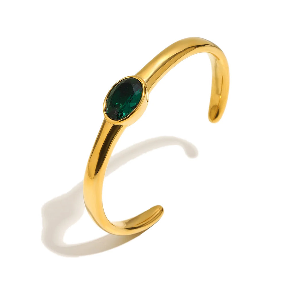 Gold oval stone open cuff bracelet in emerald green on white background, smooth high-polish band with deep green bezel-set oval stone at center, open back visible, long warm shadow
