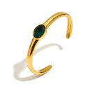 Gold oval stone open cuff bracelet in emerald green on white background, smooth high-polish band with deep green bezel-set oval stone at center, open back visible, long warm shadow
