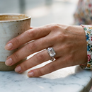 Woman's hand wearing silver emerald cut stone bezel ring resting beside ceramic cup on marble surface, large faceted stone in cool daylight, floral print sleeve, blurred outdoor cafe background

