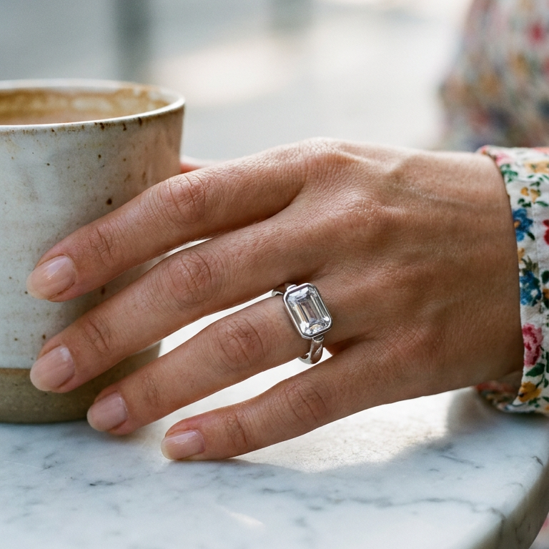 Woman's hand wearing silver emerald cut stone bezel ring resting beside ceramic cup on marble surface, large faceted stone in cool daylight, floral print sleeve, blurred outdoor cafe background
