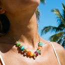 Close-up of gold colorful resin bead station necklace worn at collarbone, multicolor resin beads and polished gold spheres catching bright tropical sunlight, white tank top strap, blue sky and palm trees in background
