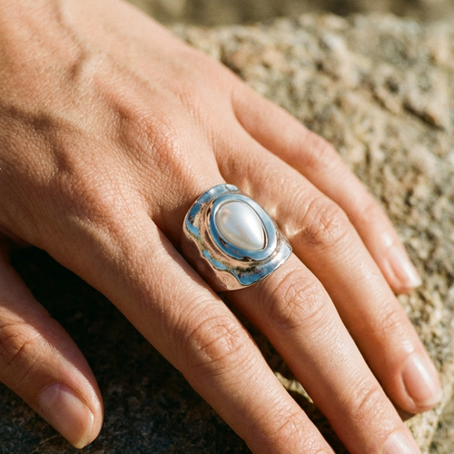 Hand wearing silver hammered pearl chunky ring resting on rough coastal rock, wide organic silver band with bezel-set teardrop pearl in bright sunlight, blurred blue sky in background
