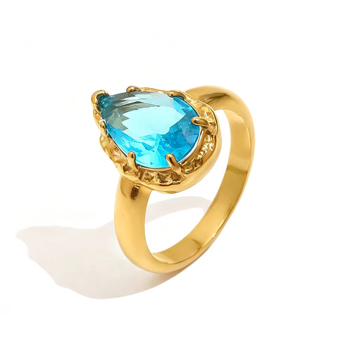 Gold blue teardrop stone halo cocktail ring on white background, aqua blue faceted teardrop stone in multi-prong ornate gold halo setting, tapered gold band, long warm shadow
