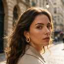 Woman wearing gold twisted frame tiger's eye stud earrings, three-quarter profile with dark wavy hair, beige trench coat, Paris street with Haussmann buildings in background
