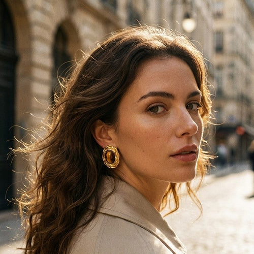 Woman wearing gold twisted frame tiger's eye stud earrings, three-quarter profile with dark wavy hair, beige trench coat, Paris street with Haussmann buildings in background
