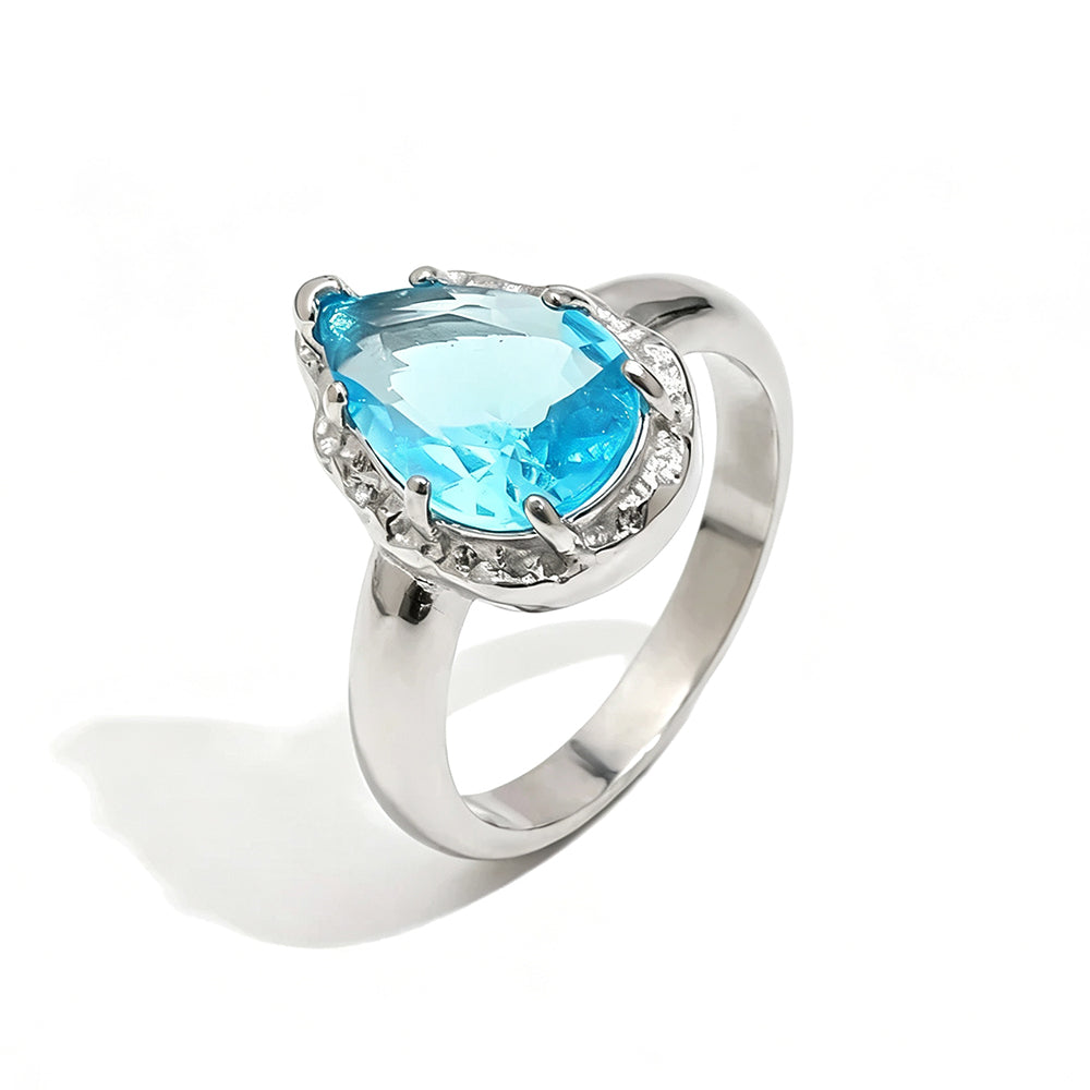 Silver blue teardrop stone halo cocktail ring on white background, ice blue faceted teardrop stone in multi-prong silver halo setting, tapered silver band, soft shadow
