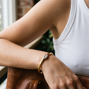 Woman wearing gold oval stone open cuff bracelet in clear, arms crossed resting on brown leather bag, white ribbed tank top, bezel-set clear oval stone visible, blurred greenery in background
