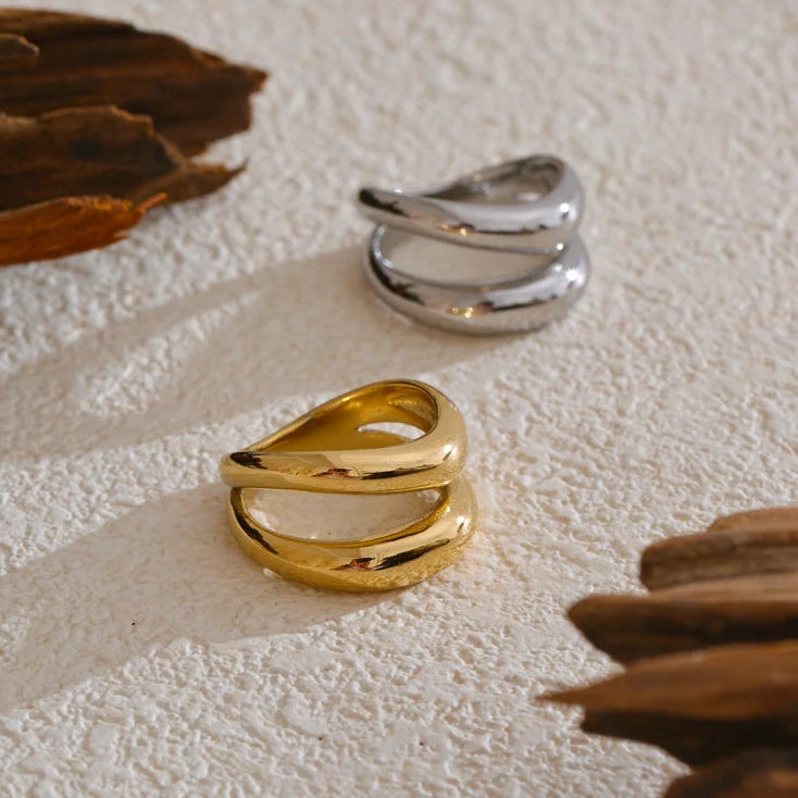 Gold and silver double wave band rings displayed on textured white stone surface, gold ring in foreground and silver ring behind showing identical two-band wave structure, dried leaves as props, warm natural light

