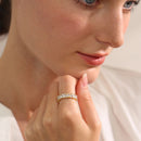 Woman's hand raised to chin wearing gold heart-cut eternity ring on middle finger, heart-shaped stones catching soft diffused indoor light, blurred neutral background, close-up portrait composition
