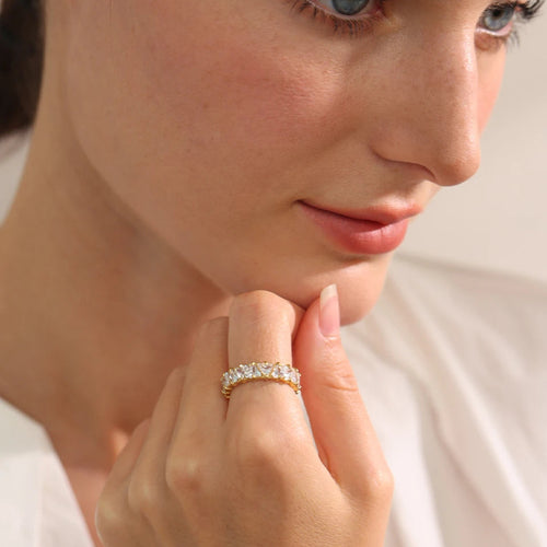 Woman's hand raised to chin wearing gold heart-cut eternity ring on middle finger, heart-shaped stones catching soft diffused indoor light, blurred neutral background, close-up portrait composition
