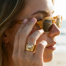 Woman wearing gold wavy wide band ring at beach, amber crystal circle on index finger, adjusting sunglasses
