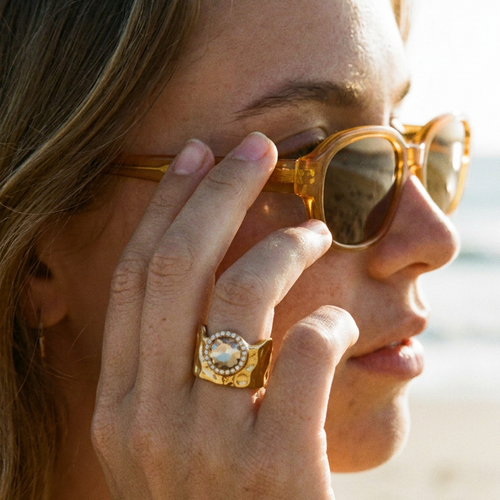 Woman wearing gold wavy wide band ring at beach, amber crystal circle on index finger, adjusting sunglasses
