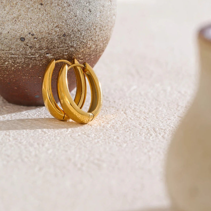 Pair of gold chunky hoop earrings leaning against dark brown ceramic sphere on cream textured stone surface, thick mirror-polished tube hoops with hinged clasps visible, warm side light casting soft shadows
