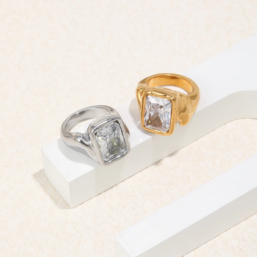 Gold and silver oversized emerald cut chunky rings resting on white plaster step against cream background, side angle showing organic band profile, chunky band thickness and irregular shoulder outline visible
