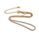 Gold round stone tennis necklace on white background, continuous prong-set round stones along full length, lobster clasp with extender chain and round tag pendant, warm light with soft shadow beneath

