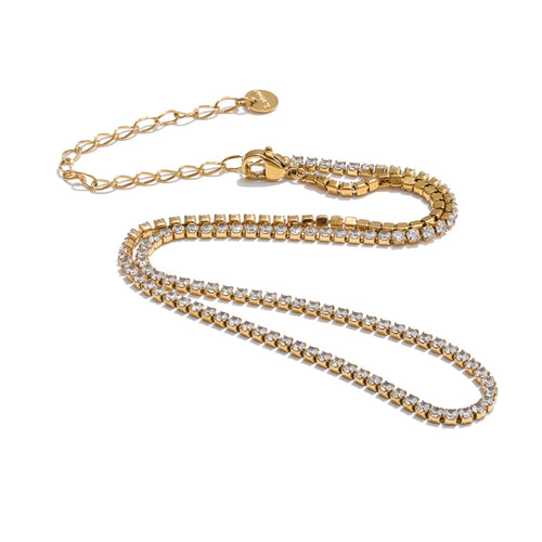 Gold round stone tennis necklace on white background, continuous prong-set round stones along full length, lobster clasp with extender chain and round tag pendant, warm light with soft shadow beneath
