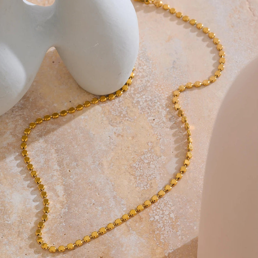 Gold disc chain necklace laid loosely on pink marble surface, flat disc links showing round polished faces and thin edges in warm side light, white ceramic vase in upper left corner, warm blush stone background

