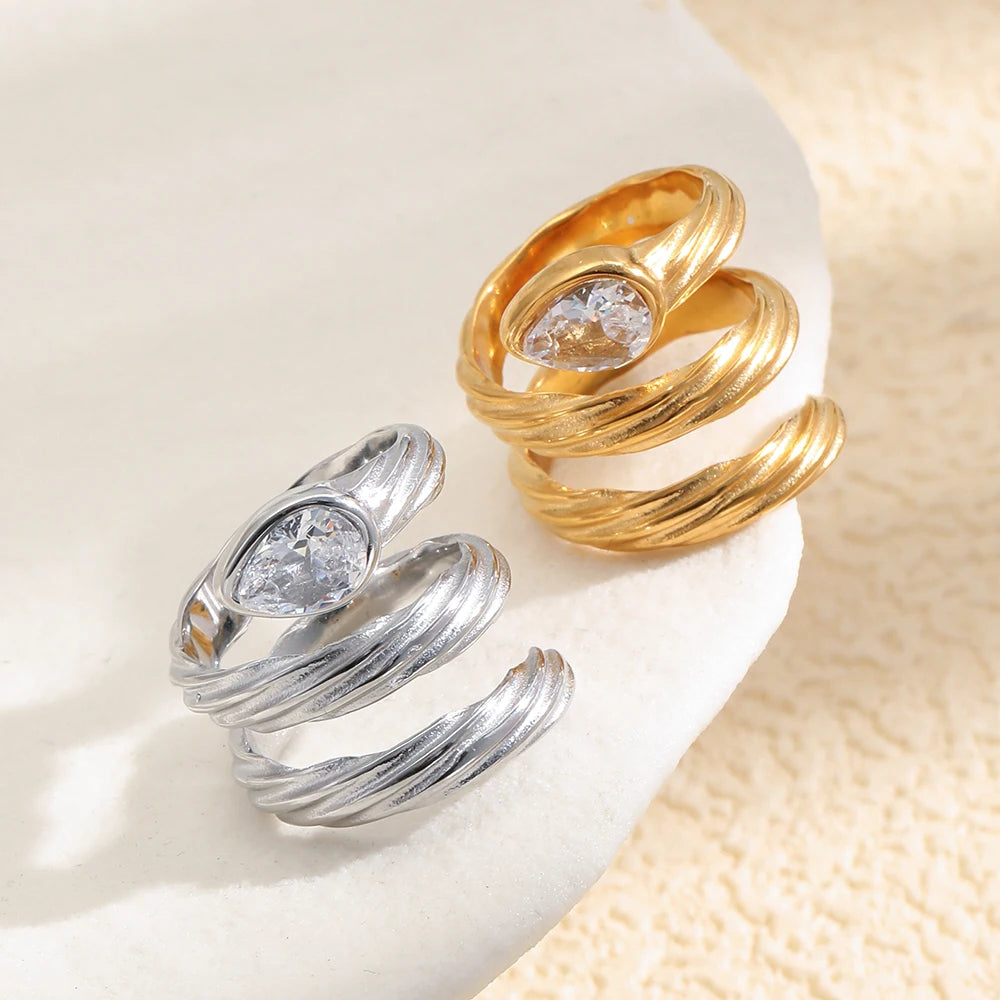 Gold and silver spiral wrap rings on white stone surface, textured coiling band and pear-cut crystal detail visible
