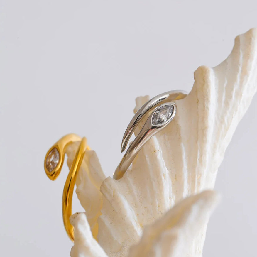 Gold and silver marquise stone snake rings displayed on white coral branch against light grey background, both rings showing bezel-set marquise stones at snake head and open adjustable band, soft diffused light
