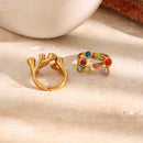 Gold multicolor stone scatter open band ring and plain gold open band ring side by side on cream stone surface, nine colored bezel-set stones visible, white ceramic object in background
