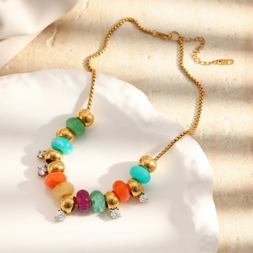 Gold colorful resin bead station necklace laid on white coral stone surface, multicolor resin beads and polished gold spheres with clear stone stations, box chain and lobster clasp with extender visible, soft white fabric in background
