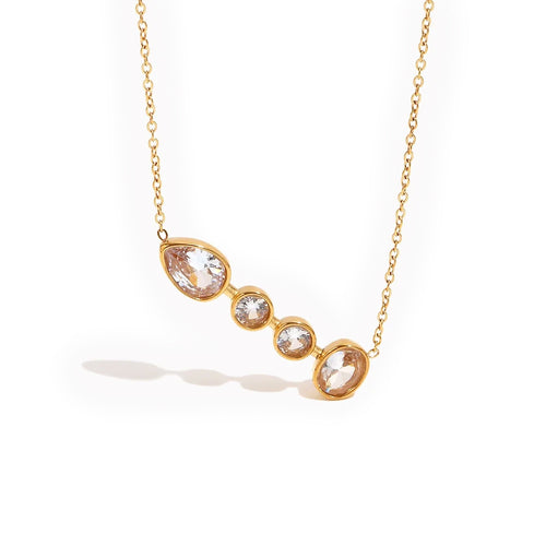 Gold graduated bezel stone bar necklace on white background, angled view showing teardrop and three round bezel-set clear stones, fine cable chain, soft shadow
