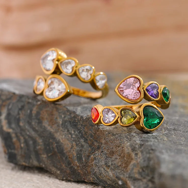 Gold white and multicolor heart stone open rings displayed on rough stone surface, white ring in background showing clear heart stones, colorful ring in foreground showing pink, purple, green, red and olive heart-cut stones, warm wood background
