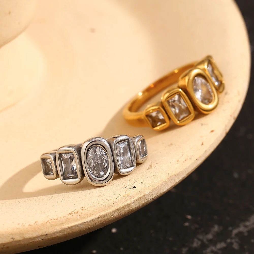 Gold and silver oval and cushion multi-stone rings resting inside cream ceramic dish, both colorways showing bezel-set oval center stone and four cushion accent stones, warm cream and dark marble background
