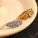 Gold and silver oval and cushion multi-stone rings resting inside cream ceramic dish, both colorways showing bezel-set oval center stone and four cushion accent stones, warm cream and dark marble background
