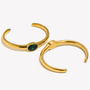 Gold oval stone open cuff bracelets showing front of emerald green version and back of clear version, open cuff structure and smooth band arc visible, white background with warm directional shadow
