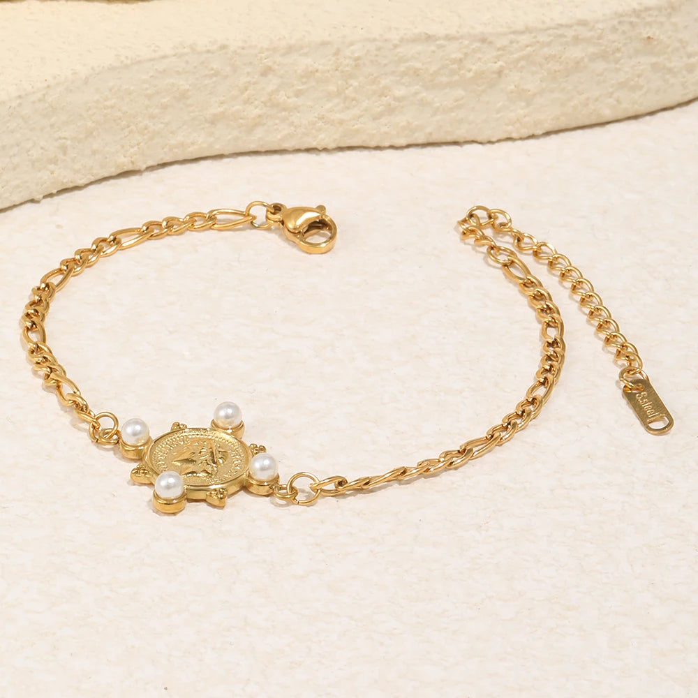 Gold pearl coin charm Figaro chain bracelet laid on cream stone surface, engraved coin charm with four pearl accents, alternating Figaro chain links, lobster clasp extender and branded tag visible, warm directional light

