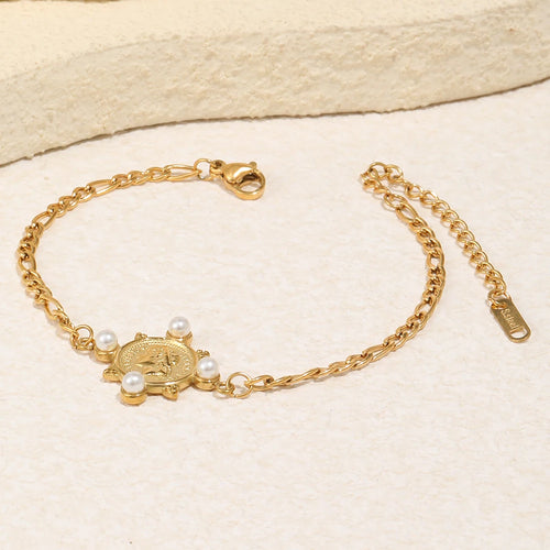 Gold pearl coin charm Figaro chain bracelet laid on cream stone surface, engraved coin charm with four pearl accents, alternating Figaro chain links, lobster clasp extender and branded tag visible, warm directional light
