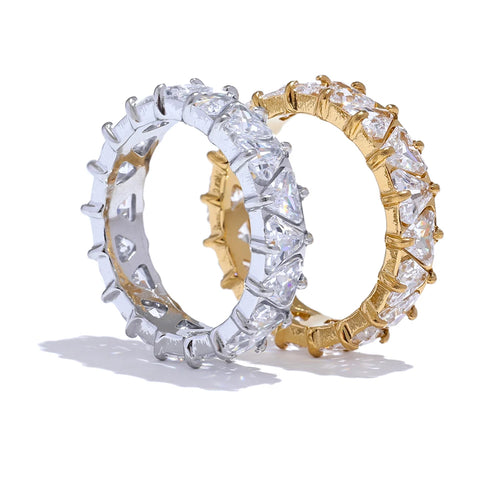 Gold and silver heart-cut eternity rings standing upright on white background, full band of heart-shaped stones in four-prong settings around both rings, warm and cool metal colorways shown side by side, long soft shadows
