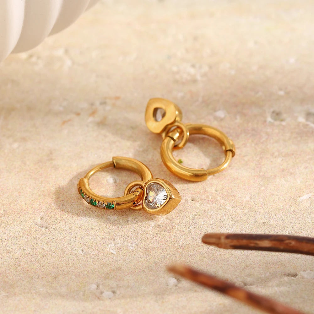 Two gold green stone pavé heart charm hoop earrings on white marble surface, one showing pavé hoop face and heart charm, one showing back, tortoiseshell hair clip nearby
