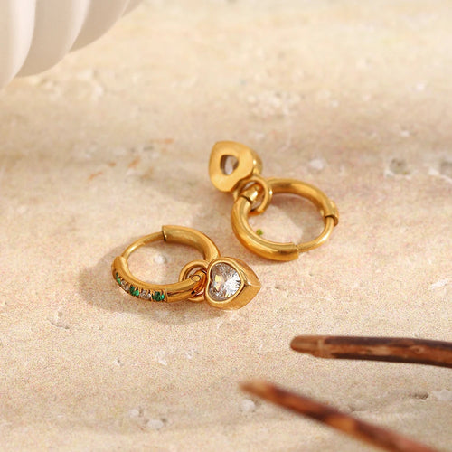 Two gold green stone pavé heart charm hoop earrings on white marble surface, one showing pavé hoop face and heart charm, one showing back, tortoiseshell hair clip nearby
