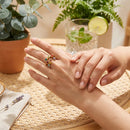 Woman wearing gold multicolor stone scatter open band ring on middle finger, hands resting on woven rattan surface, green plants and lime water glass in background
