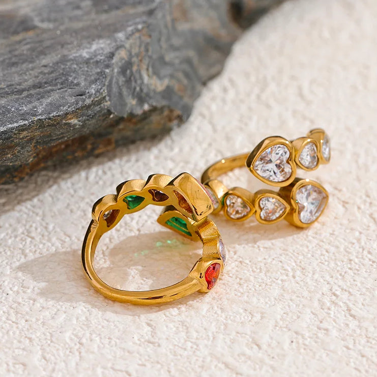 Gold heart stone open rings displayed from back angle on white sandy stone surface, colorful ring on left showing green and red stones from reverse, white ring on right showing clear heart stones, open band gap visible on both, grey rock in background
